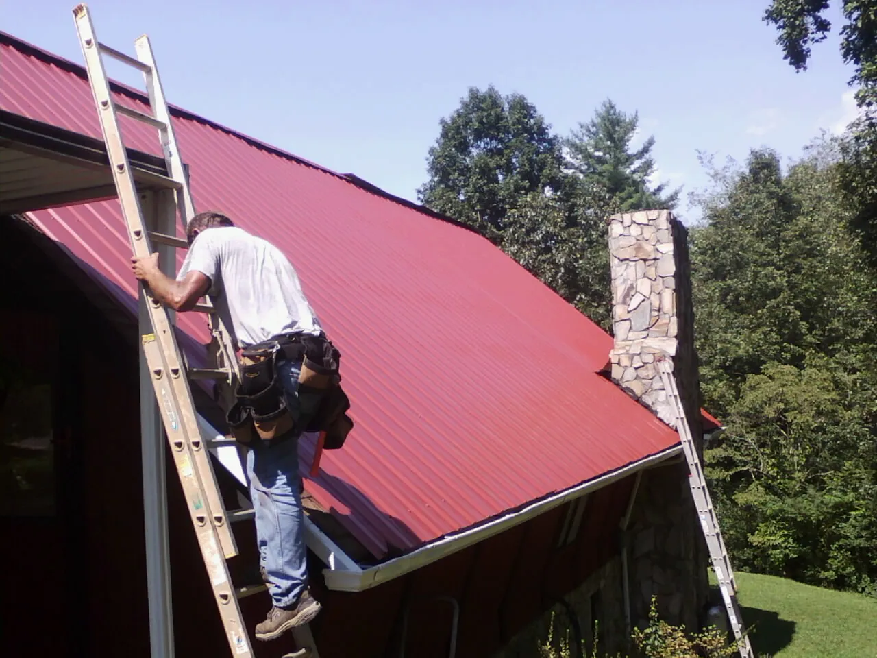 Roofer on a ladder beside a red metal roof and stone chimney — Soffit & Fascia Repair by Roofing Brothers in Winfred